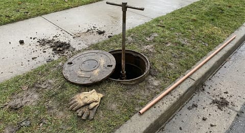 An open curb stop box in the grass strip by a sidewalk with a T-handle valve key in the shaft and new copper tubing lying at the curb.